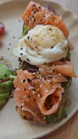 A close-up, top-down shot shows a dish of food. In the foreground is a slice of toast topped with smoked salmon, greens, and a poached egg, sprinkled with sesame seeds. To the left, sliced avocado is drizzled with a dressing and sprinkled with sesame seeds. Further to the left, halved cherry tomatoes are also dressed and sprinkled. The background is blurred but appears to be a table with other dishes and drinks. The lighting is natural and the focus is on the food.