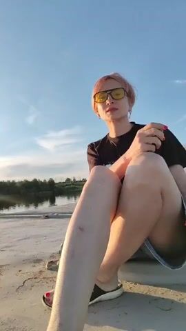 A woman with short, pinkish-blonde hair and yellow sunglasses is sitting on a large, flat, metallic surface. She is wearing a black t-shirt with a graphic design and denim shorts. Her legs are crossed, and she is looking towards the camera with a slight smile. She has red nail polish on her toes, visible through her sandals.

Behind her, there is a large, white building with multiple windows, which appears to be part of a ship or a large industrial structure. To her left, there is a body of water, likely a river, with greenery along its bank. The sky is clear with some wispy clouds, and the lighting suggests it is late afternoon or early evening, with the sun casting long shadows. The metallic surface she is sitting on has visible rivets and circular hatches. The overall scene appears to be set in an industrial or port area. No visible text is present in the media.