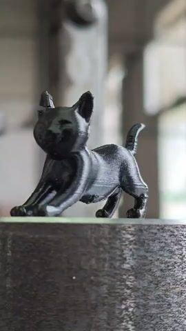 A small, black, 3D-printed cat figurine is positioned on a textured gray surface. The figurine is in a stretched-out pose, with its front paws extended forward. The background is blurred, but appears to be an indoor setting with some industrial or machinery elements visible, and possibly natural light coming from a window or opening. The camera is focused on the figurine, which is the primary subject of the video.