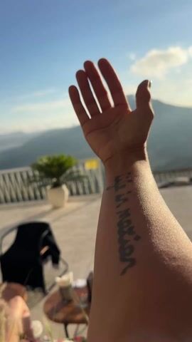 The video shows a person's arm with a Greek tattoo held up against a backdrop of a scenic mountain view. The arm is fair-skinned and the tattoo is in black ink, running vertically along the forearm. The hand is outstretched with fingers slightly splayed. In the background, there is a patio or terrace with a low white railing. A small green plant in a white pot sits behind the railing. A small wooden table with some items on it, including a smartphone and a beverage, is also visible. There is a black shawl draped over a chair. The mountain range is the prominent feature, with the peak of the mountain partially obscured by clouds. The sky is blue with scattered clouds. The lighting suggests it is daytime.