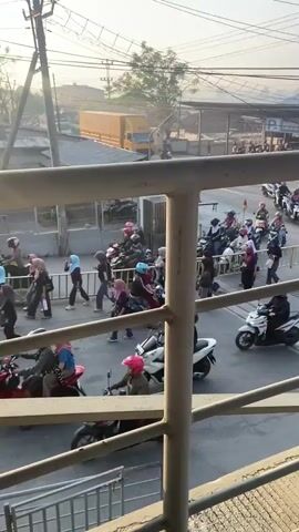 The video captures a busy street scene from a slightly elevated perspective, looking down through a metal railing. A large number of motorcycles and scooters, many with riders wearing helmets and some with passengers, are navigating the road. There are also pedestrians present, walking along the side of the road and near the motorcycles.

The environment appears to be urban, with a paved road, some overhead wires, and buildings or structures in the background. A yellow truck is visible in the distance. There are barriers or gates set up on the road, possibly indicating a crossing or a controlled area. The lighting suggests it is daytime, possibly late afternoon or early morning, with a soft, warm light. The overall impression is one of active movement and daily life in a populated area.
