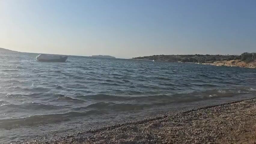 The video shows a body of water with small waves lapping at a pebble beach in the foreground. A small white boat is visible in the water to the left. The sky is clear and blue, and the sun appears to be low in the sky, casting a soft light. The shoreline in the distance is lined with trees and buildings.