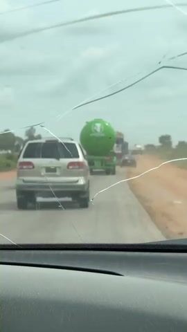 The video shows a view through a cracked windshield of a vehicle traveling on a paved road. The road has a dirt shoulder on the right side. In front of the vehicle, there is a silver SUV, followed by a green tanker truck with a white logo. Several other vehicles are visible further down the road. The sky is overcast. The interior of the vehicle, including the dashboard, is visible in the foreground.