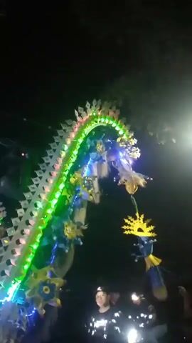 The video shows a nighttime outdoor scene with a decorative illuminated dragon-like structure as the main focus. The structure is adorned with colorful LED lights that change in sequence, predominantly blue, pink, and white. It appears to be part of a parade or festival float, with what looks like a large speaker system and other lighting equipment positioned to its left. In the foreground, several people are visible, their faces mostly obscured by the darkness and the bright lights. The ground is paved, suggesting a street or road. The overall atmosphere is festive and lively, typical of a nighttime celebration.