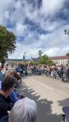 The video shows a blue vintage car driving along a road, past a large crowd of spectators on both sides. The car, a Bugatti Type 35, is driven by a man who waves to the crowd. The crowd is behind metal barricades. Many people are holding up their phones to record the event. The buildings and architecture in the background are European in style. The sky is blue with scattered clouds. The video seems to have been taken at an outdoor event in a European town, where a vintage car parade is taking place. The angle is slightly from below the heads of the people closest to the camera, emphasizing the height of the car and crowd.