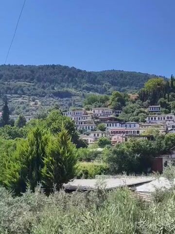 A drone shot pans across a hillside village. The village is composed of white houses with red tile roofs nestled among lush green trees. The houses are clustered together and appear to be built on terraces. The background features a steep, forested mountain under a clear blue sky. In the foreground, there are dense bushes and trees obscuring the view of some buildings. The overall impression is of a serene and picturesque rural landscape.