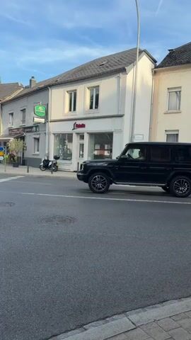 A black Mercedes G-Wagon is driving through an intersection. The car is moving from left to right in the frame. There are buildings on either side of the street, and a traffic light is visible in the background. The sky is blue with some clouds. A person is visible in the driver's seat of the car.