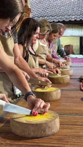 In a well-lit indoor setting, six individuals are participating in what appears to be a cooking class. They are lined up at a long wooden table, each equipped with a round, wooden cutting board and a large, silver knife. The participants are all wearing tan aprons, suggesting a unified activity. They are focused on chopping yellow, waxy-looking ingredients, possibly garlic or ginger. The woman closest to the camera has bright red nail polish. The table is made of a woven material, and the background shows glimpses of a tiled roof and other decorative elements, suggesting a location in Southeast Asia.