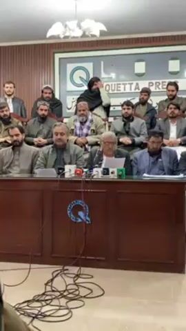 The video shows a press conference, likely in Quetta, Pakistan. A group of men are seated behind a long, wooden table. The table is cluttered with microphones and wires. One man in the center is speaking from prepared notes. Behind the table, a sign reads "QUETTA PRESS CLUB." A chandelier hangs from the ceiling. The men behind the table are dressed in various styles of traditional Pakistani clothing, including shalwar kameez and some are wearing head coverings. The room has wood paneling. Some of the men are looking at the speaker, while others look straight ahead or to the side.