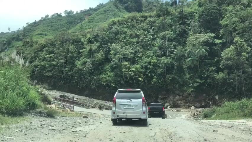 A silver minivan and a black SUV are driving on a rough, dirt road in a mountainous, tropical region. The minivan is in the foreground, followed by the SUV. The road is lined with lush green vegetation and trees. There is a partially constructed bridge or roadwork on the left side of the road. The sky is overcast. The video appears to be shot from inside a moving vehicle.