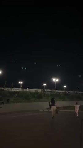 This video appears to be filmed at night in an outdoor public space, possibly a park or promenade, in Kaspiysk, Russia. The scene is dimly lit by numerous streetlights casting a warm glow on a paved walkway and surrounding greenery. Several people are visible, including a woman with dark hair walking away from the camera, a young child in a light-colored dress, and a person in a red shirt. In the distance, more figures can be seen strolling along the illuminated path.

The sky is dark, and there are several faint, bright lights scattered across it, some appearing as pairs of glowing orbs. These could be distant lights, reflections, or possibly even astronomical phenomena. The overall atmosphere is serene, suggesting a peaceful evening outing. A tall, white pole, possibly a flagpole or part of a light fixture, stands prominently on the right side of the frame. Beyond the park area, some buildings and scaffolding are visible, along with what seems to be smoke or mist rising, illuminated by lights. The time of day is clearly nighttime, and no weather conditions are particularly apparent beyond the clear, dark sky. No text is visible in the video.