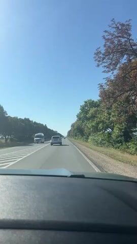 This is a first-person perspective video, likely from inside a vehicle, looking forward. The road ahead is paved, with multiple lanes and white lane markings. Several cars are visible on the road, including a white van in the left lane and a white car in the right lane. Trees and foliage line both sides of the road, with a clear blue sky overhead. The vehicle appears to be in motion, traveling down the highway.