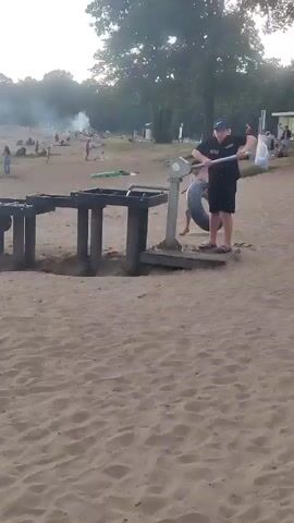 The video shows a sunny day at a sandy beach next to a lake. Several people are enjoying the water and the beach. In the foreground, a group of children are playing around a water table. The water table is made of dark material and has several compartments that are likely filled with water. One boy in a white t-shirt and dark shorts is pushing a ball around in one of the compartments. Another boy in a blue t-shirt and dark shorts is standing next to him, watching. A third child is partially visible behind the water table.

In the background, there are more people on the beach and in the lake. Some are swimming, while others are sitting or walking along the shore. A plume of smoke rises in the distance, possibly from a barbecue or campfire. The trees in the background suggest a natural, park-like setting. The overall atmosphere appears to be relaxed and enjoyable, typical of a summer day at the beach. The lighting suggests it is daytime, likely late afternoon or early evening, given the soft light and long shadows. There is no visible text in the video.