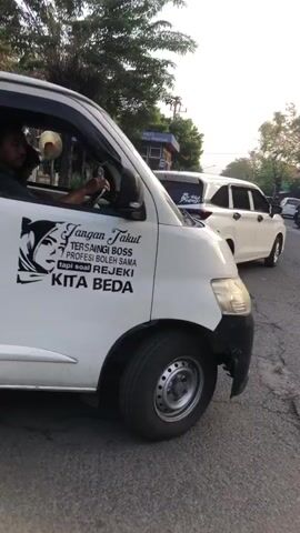 The video shows the side view of a white pickup truck parked on a paved surface. The truck has a decal on its door that says "JANGAN TAKUT TERSEMAINGI BOSS TAKUT TERSEMAINGI SAMA KITA BEDA REJEKI KITA BEDA" which translates to "DON'T BE AFRAID OF BEING ALIENATED BOSS BE AFRAID OF BEING ALIENATED FROM US, OUR BLESSINGS ARE DIFFERENT". There is a person visible inside the driver's seat. In the background, there are trees and other vehicles. The lighting suggests it is daytime.