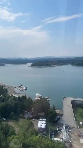 An aerial view captures a wide, calm lake surrounded by lush green forested hills under a bright blue sky with scattered white clouds. On the left side of the frame, a shoreline features a small marina with several boats docked, including a few white motorboats and a larger white vessel. Adjacent to the marina are some small buildings and trees. A dirt road winds through the trees, with a few parked vehicles visible. In the foreground, on the right side of the image, a concrete structure, possibly a dam or pier, extends into the water. A white, open-air structure with circular white coverings is visible near the edge of the water, and a tall, white metal tower, possibly a crane or observation structure, stands nearby. The overall scene suggests a recreational area at a lake on a sunny day.
