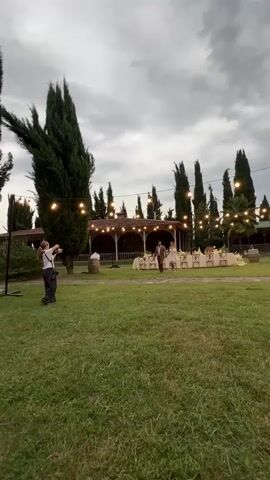 A woman stands on the left side of the frame holding a camera and aiming it toward the right. She is wearing a black skirt and a light-colored shirt. Her hair is dark and pulled back. She is standing in a grassy area. Behind her, there are tall, dark green trees. A string of lights hangs from poles in the foreground. In the midground, a couple is walking across the grass. The man is wearing a suit, and the woman is wearing a light-colored dress. Behind them is a building with a covered patio. There are tables and chairs set up on the patio, and more string lights are hanging above. The sky is cloudy.
