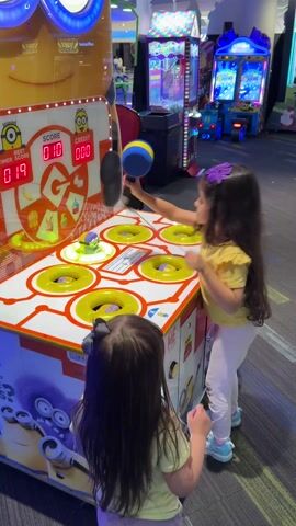 Two young girls are in an indoor arcade, which is brightly lit and features numerous colorful games. In the foreground, a girl with long dark hair and a purple hair clip, wearing a yellow shirt and light-colored pants, is actively playing a Minions-themed arcade game. She holds a blue and yellow hammer and is enthusiastically hitting the pop-up Minion figures in six circular holes on the game's surface. The game's screen displays "SCORE 011" (which changes to 012), "CREDIT 000", and "TIMER SCORE 018" (counting down). Minions characters are depicted on the base of the machine.

Another younger girl, with dark hair tied back with a gray bow and wearing a light yellow top, stands slightly behind and to the left of the player, watching the game intently. In the background, other arcade machines are visible, including claw machines with "CANDY CRANE" text and a racing game. The floor is carpeted with a patterned design. The overall atmosphere is lively and engaging for children. The location is in Columbus, United States of America.