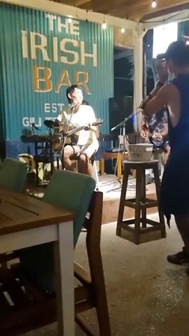 The video shows a man dancing in front of a band performing at an outdoor bar. The band consists of a guitarist, a woman singing and playing an instrument, and a keyboardist. The man dancing is wearing a black tank top, a black skirt, a red fanny pack, a baseball cap, and black sneakers. He is making various movements and gestures as he dances. The bar is decorated with string lights, and the sign behind the band reads "The Irish Bar EST." There are tables and chairs in front of the stage. A sign in the background advertises free shots, a free shirt, and a free bucket.