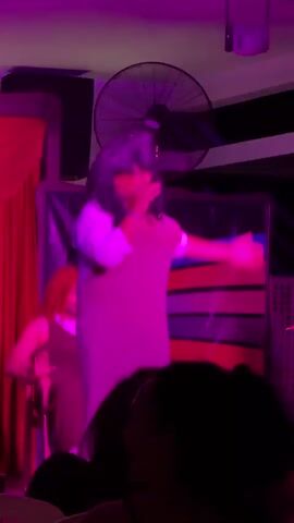 The video shows a person singing into a microphone on a stage, illuminated by blue and red lights. The singer is wearing a light-colored dress and has long, dark hair. To the left of the singer, another person is visible, wearing a dark top and what appears to be a wig. Behind them, a large fan is mounted on the wall, and red curtains hang on the left side of the stage. The audience is partially visible in the foreground, with their heads and shoulders showing. The overall atmosphere appears to be a performance or entertainment event in a dimly lit venue.