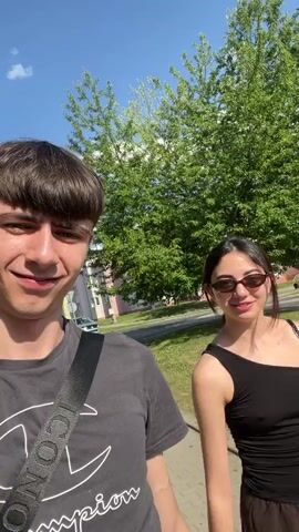 This media file captures a brief, informal moment between two young people outdoors. The primary subjects are a young man and a young woman, both appearing to be in their late teens or early twenties. The man, in the foreground and closer to the camera, wears a dark grey t-shirt with a white Champion logo and text. He has dark, short hair styled in a bowl cut and is smiling broadly, at one point making a wide, humorous grimace. A dark strap, possibly from a bag, crosses his chest.

The young woman is positioned slightly behind and to the right of the man. She has dark hair and is wearing a black, one-shoulder top and sunglasses. She smiles subtly at the camera.

The scene appears to be a daytime setting, with bright sunlight illuminating the subjects and casting shadows. The weather seems pleasant, with a clear blue sky and lush green trees visible in the background. A building with a tiled roof is also partially visible. The presence of trees and a paved walkway suggests an outdoor urban or suburban environment.

The activity appears to be casual, possibly a walk or simply filming a short video clip. The man's direct gaze and exaggerated expressions suggest he is interacting with the camera, likely recording a vlog or social media content. The overall mood is lighthearted and cheerful, conveyed through the smiles and the sunny weather.

Visible text includes the "Champion" logo and accompanying text on the man's t-shirt. The location context provided is Dobřany, Czechia, which aligns with the visual cues of a European setting.