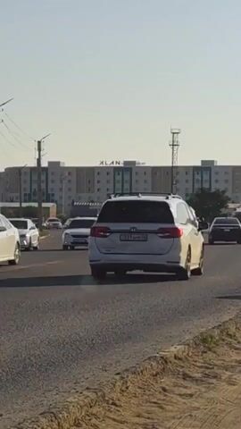 The video shows a view of a street in Atameken, Kazakhstan. The time of day appears to be late afternoon, with a clear sky and bright sunlight casting shadows. The scene is a busy road with several cars driving in the same direction. The road is paved and appears to be a main thoroughfare. 

On the left side of the road, there are power lines and poles, as well as some vegetation. In the background, there is a large, modern-looking building with the text "ALANTA" visible on its facade. The building has many windows and appears to be multi-story. There are also some smaller structures and vehicles in the distance.

The cars are diverse in make and model, suggesting a typical urban or suburban traffic flow. The overall impression is one of a functional, everyday scene in a developing area, with clear weather and active transportation. There are no people visible in the foreground, and the focus is on the road and the vehicles. The license plates of some cars are partially visible, displaying Cyrillic characters and numbers, which aligns with the location context of Kazakhstan. One car's license plate reads "134 AHC 12", another "8188 EC", and a third "615 ALE 12".