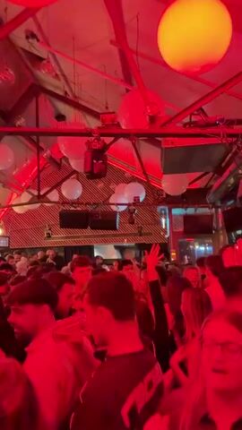 The video shows a crowded indoor setting, likely a bar or club. The space has a high, angled ceiling with exposed beams, white spherical lanterns, and disco balls. Red and white lighting fixtures are visible above the crowd. The venue is packed with people, mostly young adults, standing and talking. Some individuals are visible in the foreground, including a young man in glasses. The lighting is dim, creating a nightclub atmosphere. The background reveals wall decorations, including framed artwork and exposed lightbulbs. The camera moves slightly. The overall impression is of a lively social gathering with music and a party atmosphere.