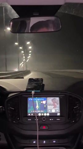 The video shows the interior of a car driving at night on a road with streetlights. The windshield is visible, showing a foggy or misty environment. A rearview mirror reflects the driver's head. A phone sits on the dashboard, and a car infotainment system displays a navigation map and music information. The emergency lights are activated.