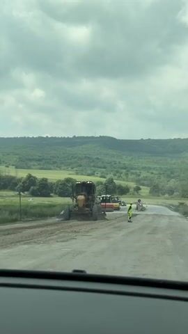 The video captures a road construction scene in Komarevo, Bulgaria, on an overcast day. In the foreground, a large yellow road grader is stationary on a dirt and gravel section of the road. To its right, a construction worker in a high-visibility yellow uniform stands, facing away from the camera, seemingly directing or monitoring the activity. Further down the road, a red and white road roller is visible, along with another construction worker. Traffic cones and barriers mark the construction area. The background features rolling green hills covered with trees under a cloudy sky, suggesting a rural or semi-rural setting. The footage is taken from inside a vehicle, with the dashboard visible at the bottom of the frame.