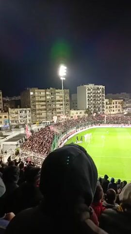 A football game is taking place in a stadium at night. The stadium is packed with fans, many of whom are wearing red and white. Flags with the same colors are waving among the crowd. The camera is positioned behind a person wearing a dark hoodie, providing an obscured view of the game. The bright floodlights illuminate the green pitch, where a few players are visible. Buildings surround the stadium, offering a glimpse of the urban environment.