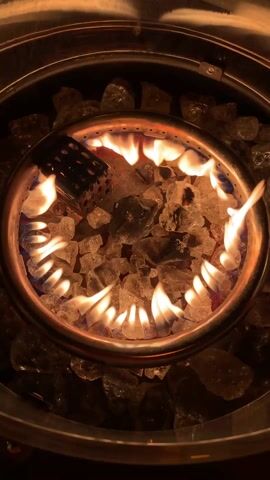 The video shows a top-down view of a lit gas fire pit.  The circular pit has a stainless steel surround, and the center is filled with clear, crystalline fire glass. Flames are coming from the center ring, and a black metal lighter is partially visible in the flame. The surrounding area of the pit is also filled with larger pieces of the same fire glass. The flames are mostly yellow-orange with hints of blue at the base. The environment is dimly lit, emphasizing the light from the flames.
