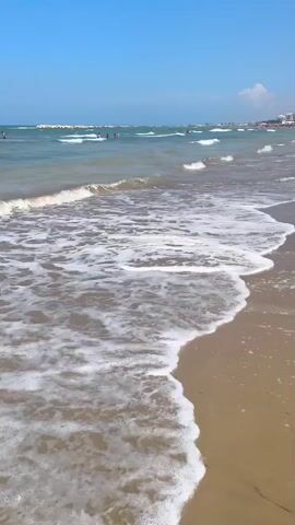 The video shows a beach scene with the camera looking out towards the ocean. The tide is coming in, with waves crashing onto the sandy shore and leaving foam. In the distance, people can be seen swimming and playing in the water. Along the shoreline, there are a few beach umbrellas and some people walking. The sky is clear and blue, indicating a sunny day. There is also a red rescue boat on the beach.