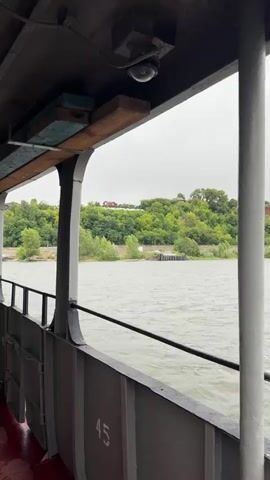 The video opens with a view from what appears to be a boat. A metal railing runs along the bottom of the frame, with a section of red flooring visible. Above the railing, vertical posts and a gray metal wall with the number 45 are visible. Above that, a wooden beam extends across, supporting a small, dark object that could be a security camera. The camera is pointed downwards. A second wooden beam is visible behind the first. The background shows a body of water, likely a river, with ripples on its surface. Across the water, a green, tree-covered hillside rises. At the top of the hill, a red-roofed house and other buildings are visible, along with large white letters spelling out a word. Several trees are on the hillside, and some smaller trees and bushes are closer to the water's edge. The sky is overcast. The video then zooms in slightly on the hillside and the lettering.