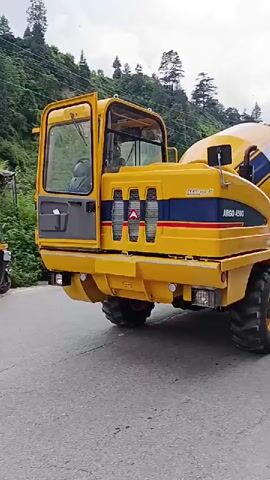 A yellow Argo 4500 cement mixer truck is shown from the side and rear. The truck is parked on a paved road with a grassy and rocky hillside in the background. Trees cover the upper part of the hill, and mountains are visible in the distance. A man wearing a black long-sleeved shirt and a light-colored baseball cap walks by the truck. The truck's cab door is open, and its drum is tilted upwards, revealing the brand name AJAX. The sky is overcast.