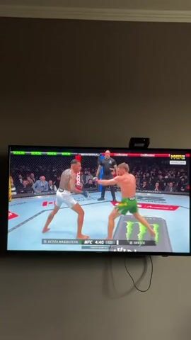 The video shows a television screen displaying a UFC fight between Della Maddalena and Prates. The fighters are in a ring, with a referee standing between them. Della Maddalena is wearing white shorts, while Prates is wearing green shorts. The fight is taking place in an arena with spectators in the background. A webcam is visible above the television.