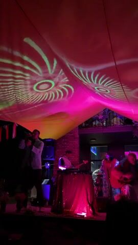 A band performs on a stage under a canopy illuminated by pink and yellow lights. Projections of circular, eye-like patterns are displayed on the canopy. From left to right, a man in a light-colored jacket plays a brass instrument and sings into a microphone. Next to him is a person behind a DJ setup, draped in a sparkling pink cloth. Further to the right, a woman in a colorful dress sings, and a person plays an acoustic guitar. A brick wall is visible behind the stage. The overall lighting is dim and atmospheric, with vibrant colored lights and projections creating a festive mood.