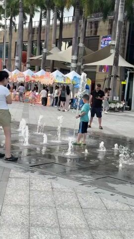 The video shows a public square in a city. The area is paved with large grey tiles. There is a water feature with multiple small fountains spraying water upwards. People, mainly adults and children, are walking, standing, and playing in the water. There are many vendor stalls covered with white and blue canopies in the background. Tall palm trees line the edge of the square. A red sculpture shaped like several interconnected trees is a central landmark. There are modern buildings and skyscrapers in the distant background, some under construction. One person is riding a small white scooter through the water. There is a Starbucks visible in the background as well. The sky is partly cloudy with the sun setting, casting a warm glow.