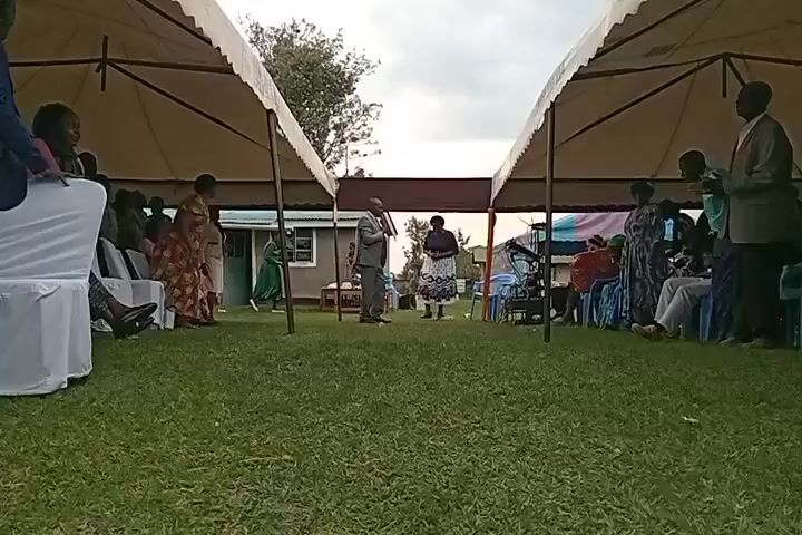 The video shows an outdoor gathering under two large tents. In the center, a man in a suit and a woman in a patterned dress are standing, facing each other. There are people sitting and standing on both sides of the grassy area, under and around the tents. Some chairs are covered in white fabric. In the background, there is a building with a green door and a small garden area. The sky is overcast. The audio consists of a person singing in Swahili.