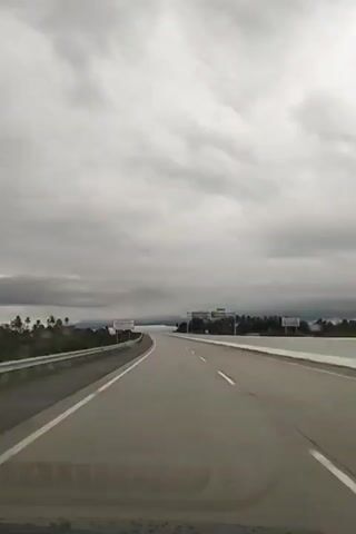 The video shows a view from inside a vehicle traveling on a highway. The sky is overcast with dark clouds. On the left side of the road, there is a metal guardrail and beyond it, a sign that appears to be in a foreign language, with text and possibly some imagery. There are also some trees in the distance on the left. The highway ahead curves gently to the right. On the right side of the highway, there are more trees and some overhead signage structures, typical of a highway system. The road itself is paved and marked with white lines. The overall impression is a drive on an empty highway under a cloudy sky.