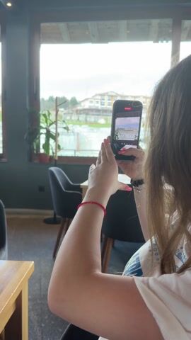 The video shows a person inside a cafe taking a video of a waterfall outside the window with their smartphone. The waterfall appears to be part of the landscape of a hotel or resort. Inside, there are tables and chairs, and a plant decoration. There is also a glimpse of an eye looking at the camera.