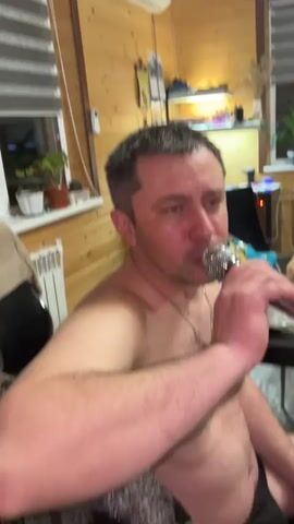 A middle-aged man with short grey hair and a silver chain is singing into a handheld microphone. He is shirtless and looks slightly emotional, with his eyes a bit red. The background shows a room with wooden walls, a window with blinds, and various items on a shelf, including plants and a lamp. There's also a glimpse of another person in the background, who appears to be sitting at a table with food. The atmosphere seems relaxed and informal.