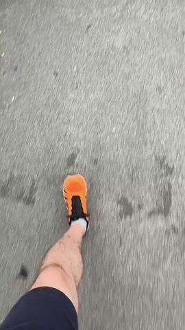 The video shows a first-person perspective of someone walking on a paved surface. The person is wearing bright orange athletic shoes with black accents and navy blue shorts. The camera angle is looking down at the person's feet and legs as they move forward. The ground appears to be asphalt or concrete, with some small debris scattered on it. The motion blur indicates the person is walking at a moderate pace.