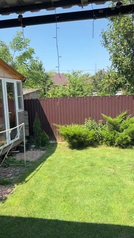 The video shows a backyard scene on a sunny day. In the foreground, a well-maintained lawn occupies most of the frame. To the left, the corner of a building with white siding and a window is visible, suggesting a house or shed. A small evergreen tree stands near the building. A brown metal fence runs across the middle ground, separating the lawn from the background. Behind the fence, several evergreen bushes are planted. Further back, trees and the roofs of houses can be seen. On the right side of the frame, a small round above-ground swimming pool with blue lining is situated on a patterned brick patio. A few small bushes are planted along the edge of the patio. The scene is viewed from under a covered patio area, with dark beams and hanging lights visible at the top of the frame.
