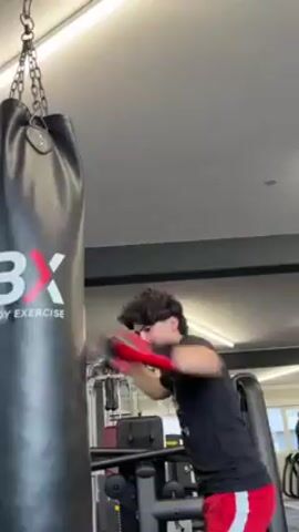 A young man with dark, curly hair is boxing at a gym. He is wearing a black t-shirt and red shorts, as well as red boxing gloves. He is punching a large, black punching bag that has the words "HBX EXERCISE HUMAN BODY EXERCISES" printed on it in white. The bag is hanging from a chain attached to the ceiling. The gym has grey walls and some weightlifting equipment in the background. The camera is positioned slightly below the man, looking up at him as he punches the bag.