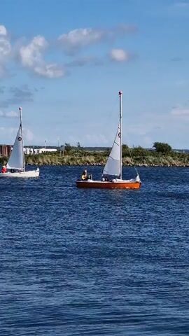 Two sailboats are on the water. The sailboat on the left is white with a white sail. The sailboat on the right is brown with a white sail. There are three people on the brown sailboat. The water is blue and there are some white clouds in the sky. There is land in the background with some trees and buildings.