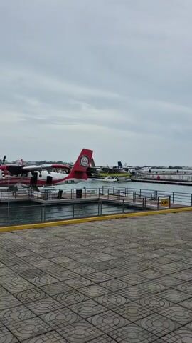 The video shows a waterfront scene with several seaplanes docked at a marina. The sky is overcast and the water is relatively calm. The foreground consists of a paved walkway with a patterned surface. A yellow curb runs along the edge of the walkway, separating it from the water. The marina includes several docks where the seaplanes are moored. The seaplanes are of various colors and designs, with the predominant color scheme being white and red. A sign with the marking "C-02" is visible on one of the docks. In the distance, a cityscape can be seen across the water. The overall setting suggests an airport or transportation hub for seaplanes in a coastal area.