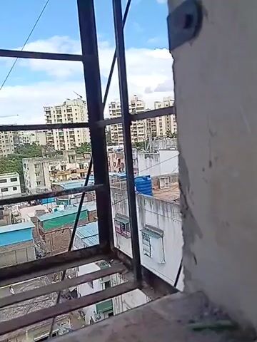 A video captures an urban panorama through a window with metal bars. The scene is a mix of residential buildings and makeshift structures. Tall, light-colored apartment buildings dominate the background, some appearing modern with many floors. In the mid-ground, there are smaller, more densely packed buildings, some with visible corrugated metal roofs in blue and green. There are also some brick structures and what appear to be smaller dwellings or shops. The sky is blue with scattered white clouds. In the foreground, the metal bars of the window frame the view. A thin wire, possibly for electricity or communication, stretches across the scene. Two figures, appearing to be small and distant, are visible on a lower rooftop or balcony, possibly engaged in activity or simply present. The overall impression is of a busy urban environment with varying levels of development.