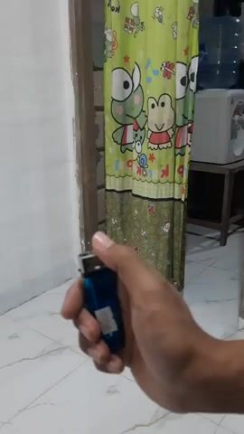 A person's hand holds a blue lighter with a flame. The background shows a wall, a doorway, a green curtain with cartoon frogs, and some appliances. The lighter is ignited and then extinguished.