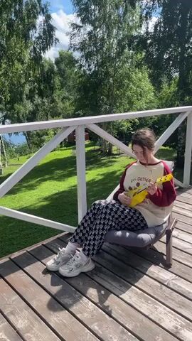A young person with dark hair pulled back is sitting in a low chair on a wooden deck, playing a yellow ukulele. They are wearing a light-colored sweatshirt with a maroon collar and sleeves, and patterned black and white pants. Their feet are in white athletic shoes. The deck has a white railing. Behind the railing, there is a green lawn and trees. Sunlight casts shadows on the deck.