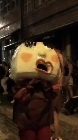 A short video shows a large puppet, resembling a person's head with blonde hair and rosy cheeks, being paraded down a street at night. The puppet's mouth is open, revealing red lips. It is attached to a person's body, who is wearing a dark, possibly red, robe. In the background, other people are visible, some watching the procession. The street is lined with buildings, and lights are visible, indicating it is nighttime. The puppet appears to be made of paper or similar material.