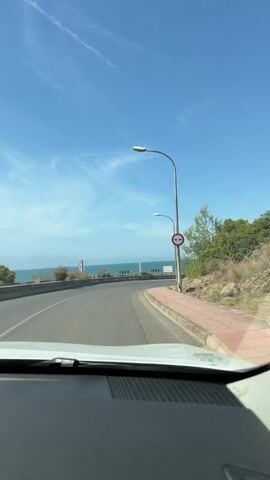 The video, shot from inside a moving vehicle, shows a picturesque coastal road with the shimmering blue sea visible on the left. The road curves to the right, leading towards a sun-drenched horizon. On the right side of the road, there's a sidewalk lined with green shrubbery and a street lamp. The bright blue sky is adorned with wispy white clouds. The vehicle appears to be moving at a moderate speed, offering a clear view of the scenic surroundings.